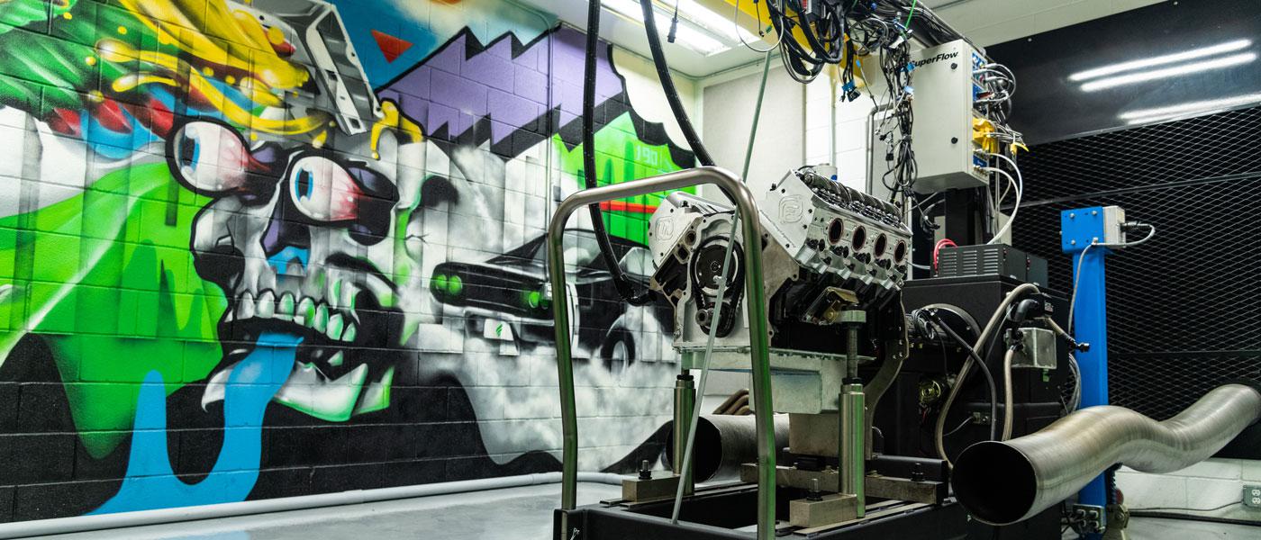 race engine hooked up to a dyno in the foreground, art mural on the wall of Frankenstein Engine Dynamics facility in the background