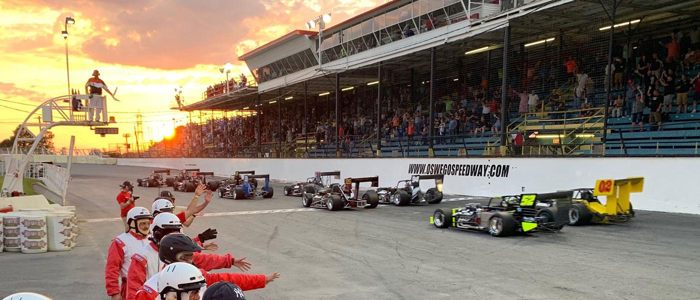 Photo of Novelis Supermodifieds at Oswego Speedway courtesy of Oswego Speedway. Packed granstands overlook the front strecth, and pit crew workers are lined up