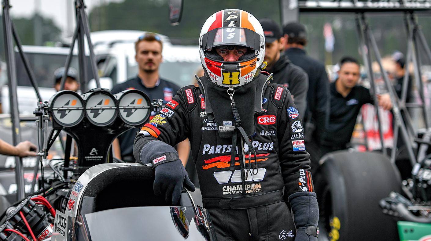 A top fuel drag racer stands outside the cockpit in his firesuit and helmet.